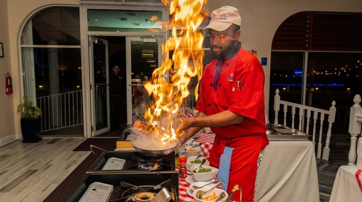 A chef performing a flambé with a pan on a table, surrounded by dining tables and chairs.