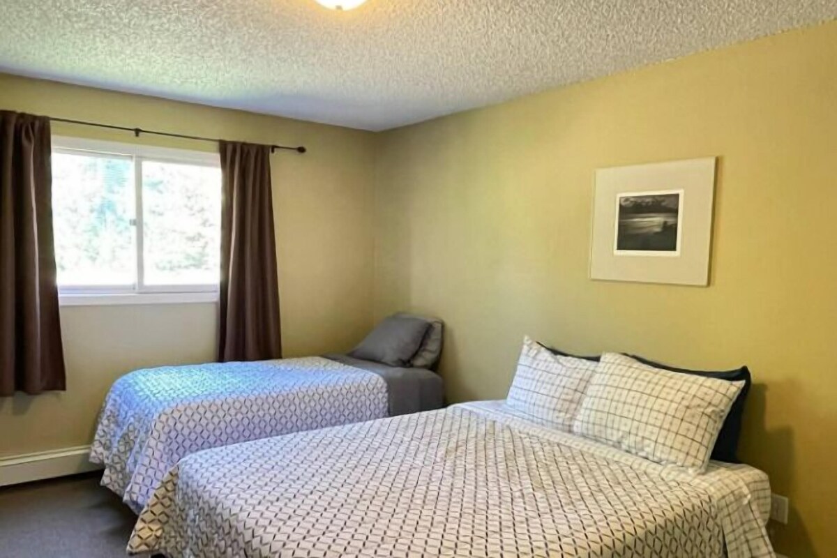 A room with two beds, a window with curtains, a framed picture on the wall, and a ceiling light.
