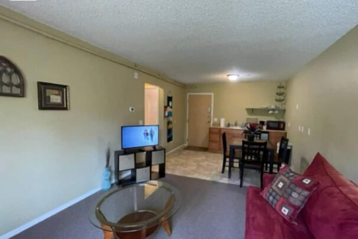 A living room with a glass coffee table, a red couch, a TV, and a kitchen area in the background.