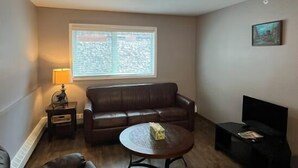 A living room with a brown leather sofa, a round coffee table, a small side table, a TV stand, and a window with blinds. - Great North Lodging (Anchorage)