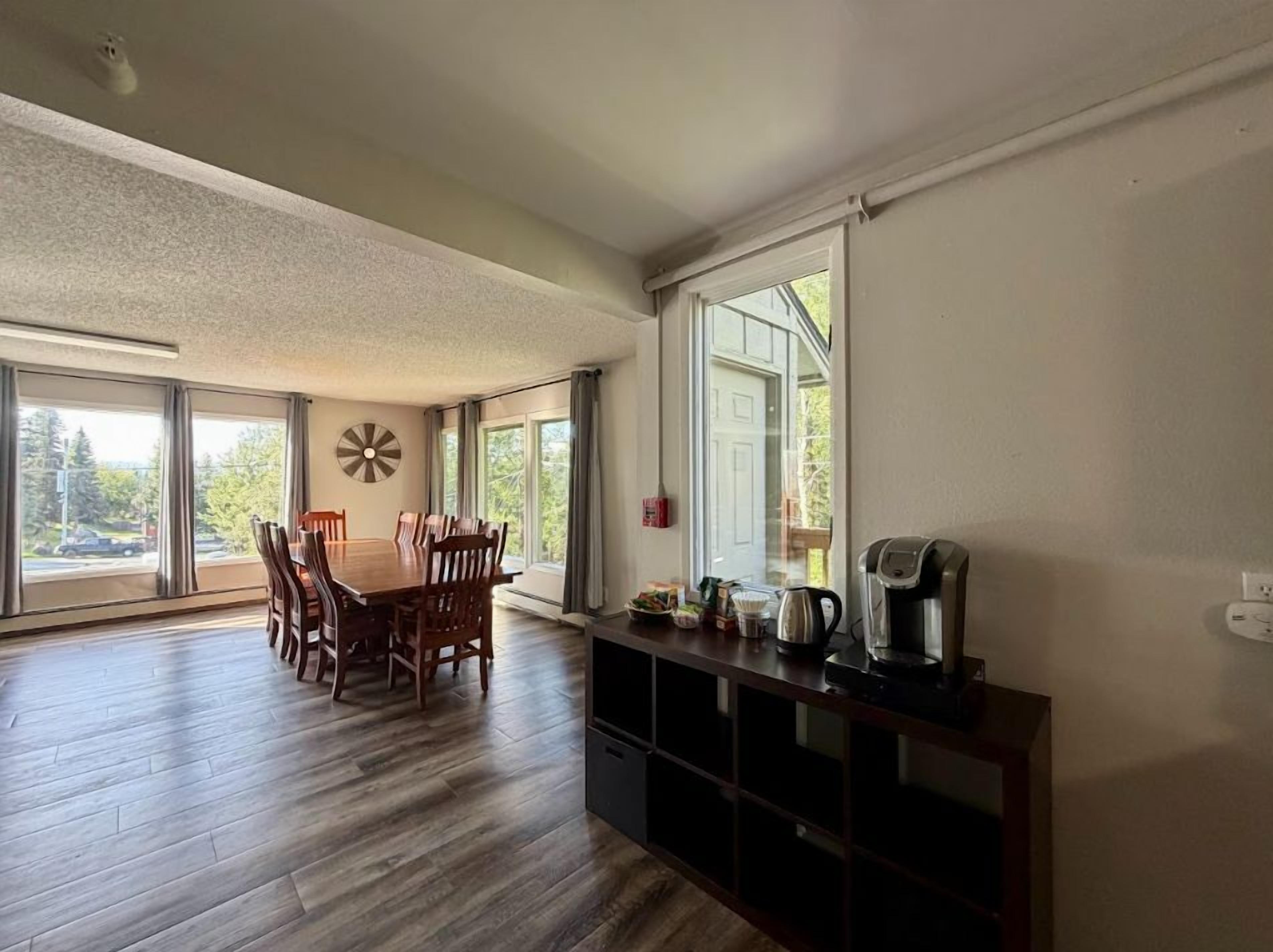 A spacious dining area with a wooden table and chairs, a coffee maker, and a view through large windows.
