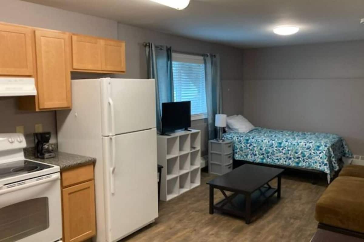 A compact kitchenette with a stove, refrigerator, and a coffee maker. There is a bed with a patterned bedspread and a small table with a lamp.