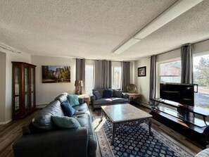 A living room with a sofa, a coffee table, and a television. There is a window with curtains, a painting on the wall, and a cabinet. - Great North Lodging (Anchorage)