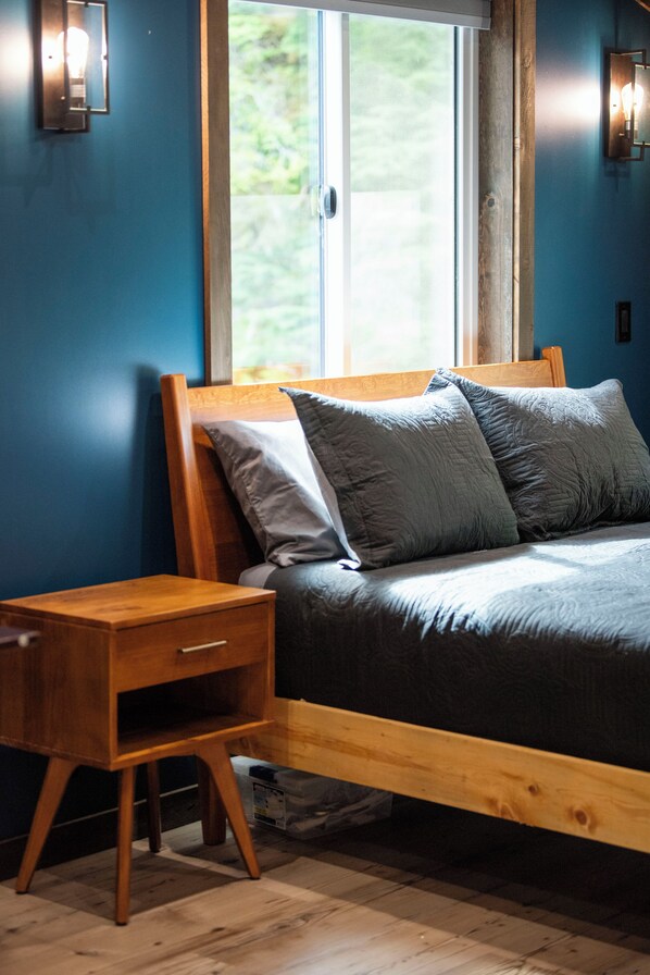 A bedroom with a wooden bed frame, a nightstand, and a window with a view of greenery. - Oz Lodge: Alice Chalet -Brand new, Alaskan chic chalet! (Seward)