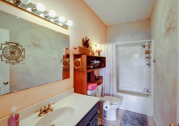 A bathroom with a white sink, a toilet, and a bathtub. There is a mirror with lights above it, and a shelf with baskets and towels. - Stylish, Eclectic two bedroom Retreat Near Downtown & Redwood (Ukiah)