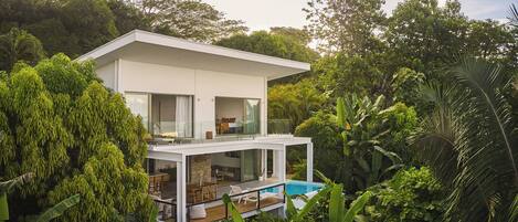 A modern two-story house with a glass balcony overlooking a swimming pool, surrounded by lush greenery.