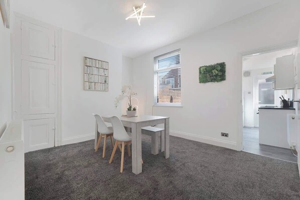 Une pièce comprenant une table à manger, des chaises, une fenêtre avec des stores, une porte et une petite plante.