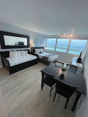 Un dormitorio moderno con una cama grande, un sofá más pequeño, una mesa de comedor con sillas y un ventanal con vistas al mar.