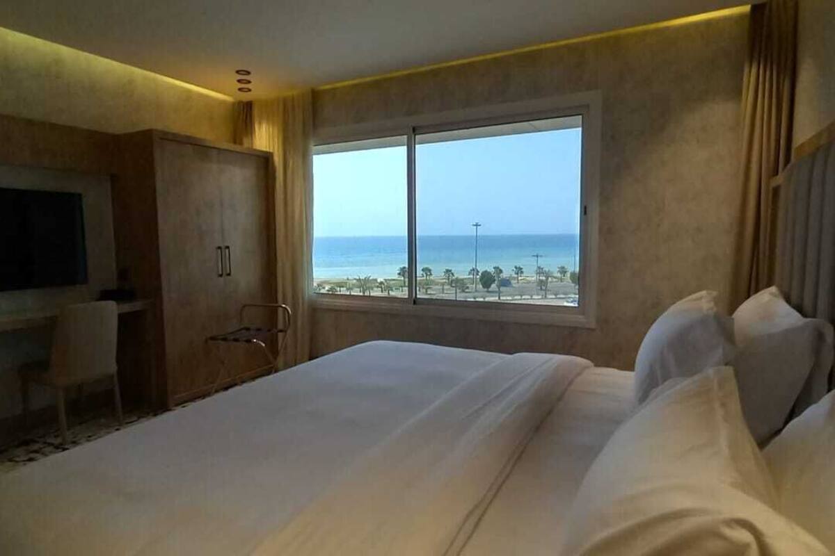 A hotel room with a large bed, a TV mounted on the wall, a wardrobe, and a window with a view of the beach and ocean.