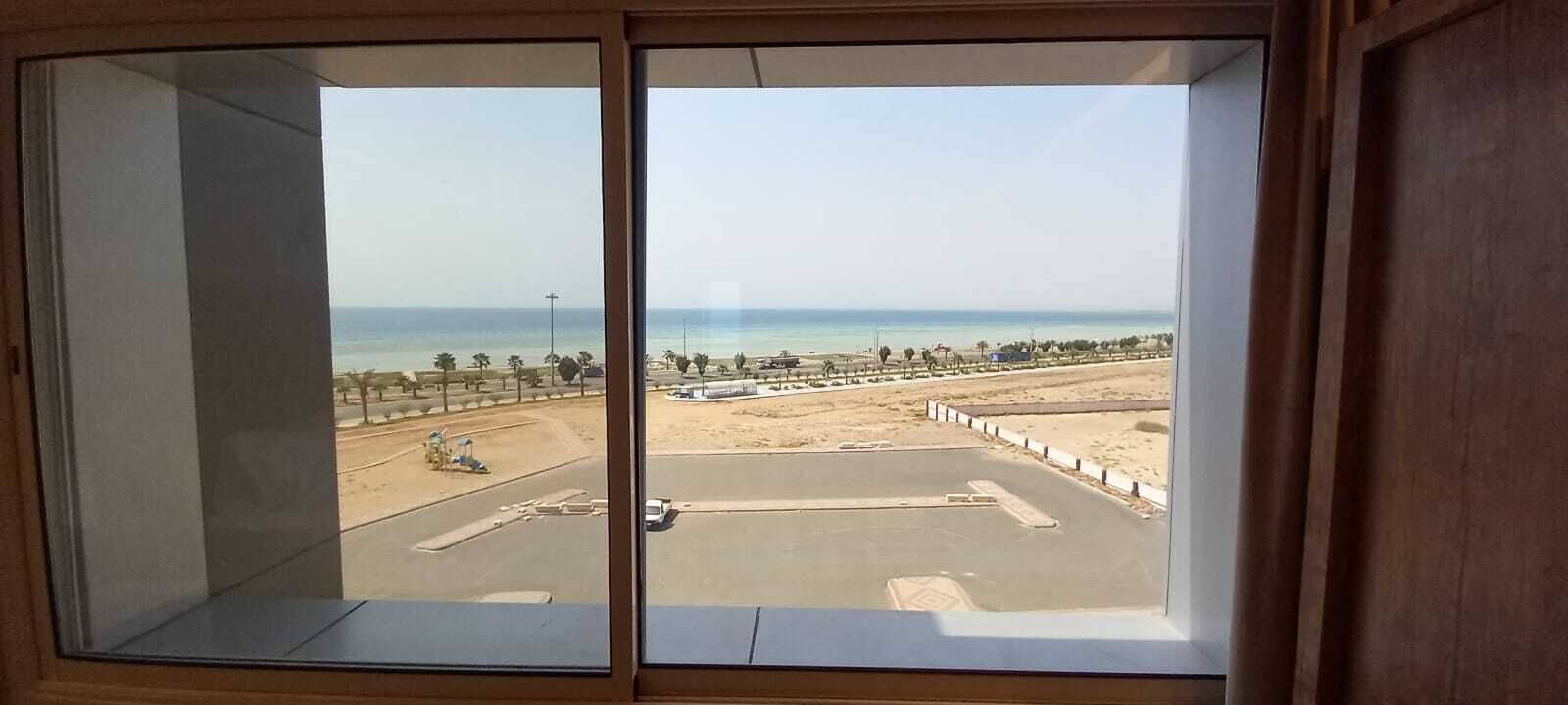 Vista des d'una finestra sobre una platja, amb una zona d'aparcament i un cel serè.