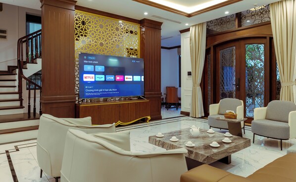A modern living room with a flat-screen TV displaying streaming services, a staircase, and a view of the outdoors. - Siêu Biệt Thự Vinhome Bason - Trung tâm thành phố (Hồ Chí Minh)