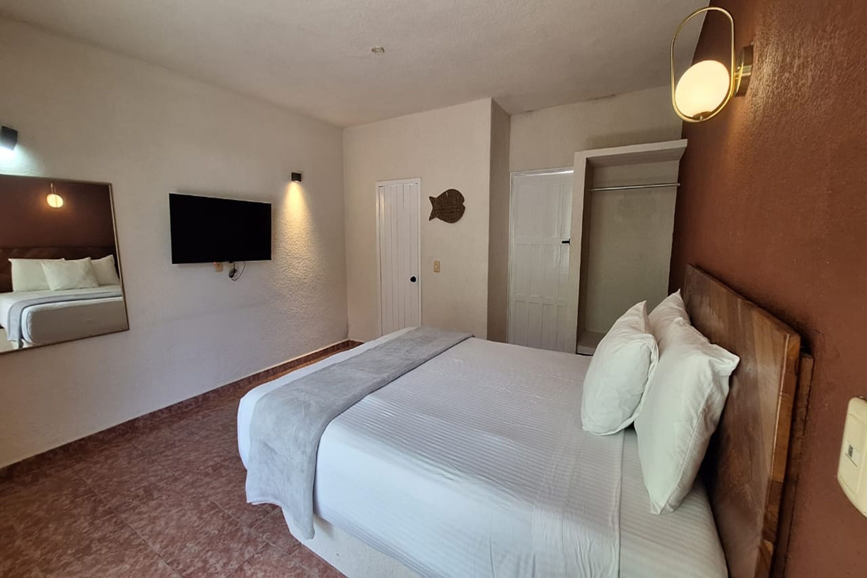 A hotel room with a large bed, a flat-screen TV mounted on the wall, a door with a fish-shaped mirror, and a light fixture above the bed.