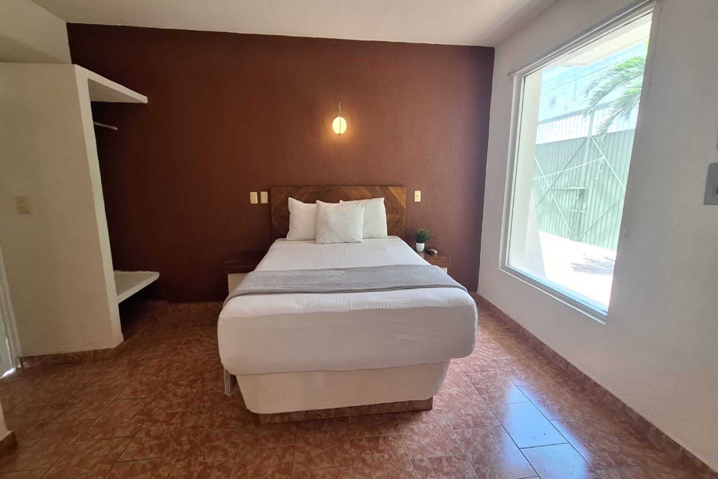 A bedroom with a large bed, a nightstand, a wall-mounted light, and a window with a view of the outside.