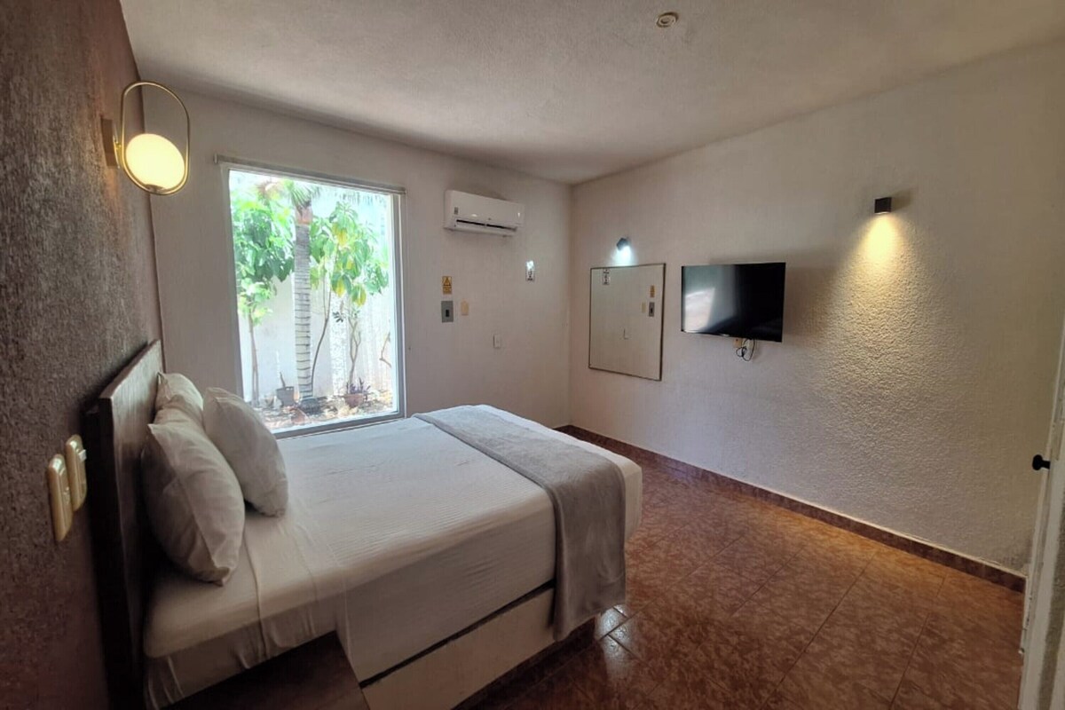 A bedroom with a bed, a window with a view, an air conditioner, and a flat-screen TV mounted on the wall.