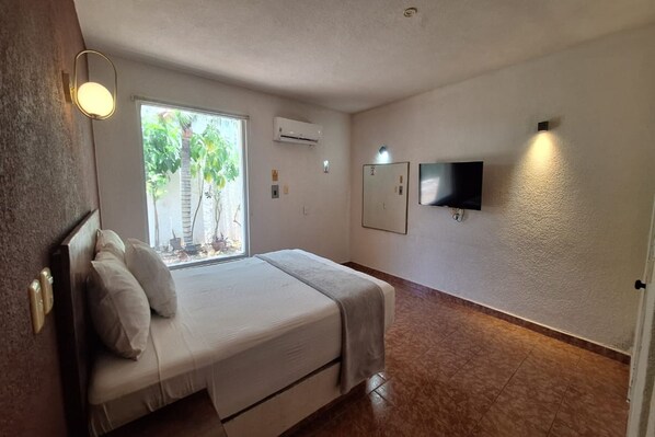 A hotel room with a bed, a window with a view of palm trees, an air conditioner, a flat-screen TV mounted on the wall, and a door. - Hotel Rolovi Cancun (Cancun)