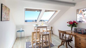 Une pièce dotée d’une lucarne, d’une table à manger avec des chaises, d’une petite table d’appoint, d’une chaise en bois et d’un vase rempli de fleurs.