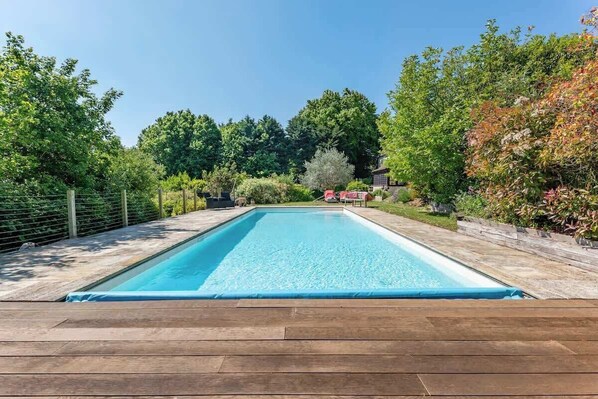 A rectangular swimming pool with a wooden deck surrounding it, set in a landscaped garden with various trees and shrubs.