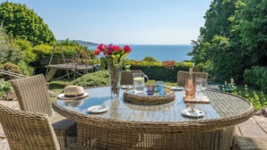Mesa redonda de mimbre dispuesta para comer, con vista al mar, rodeada de sillas de mimbre y un jarrón de vidrio con flores.