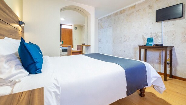 A hotel room with a bed, a wooden headboard, a television mounted on the wall, and a small table with a sign. - Hotel Mosna (Arequipa)