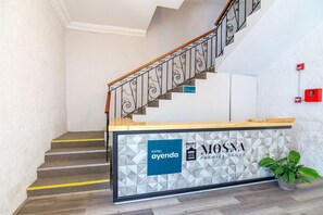 A hotel reception desk with a geometric patterned wall, a staircase with a wooden handrail, and a potted plant. - Hotel Mosna (Arequipa)