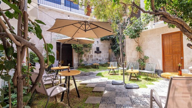 An outdoor patio with tables and chairs, surrounded by greenery and a stone wall. - Hotel Mosna (Arequipa)