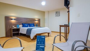 A hotel room with a bed, two chairs, a small table, a TV, and a sign with amenities. - Hotel Mosna (Arequipa)