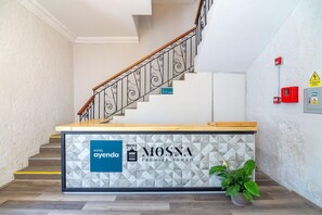 A hotel reception desk with a geometric-patterned wall, a staircase with ornate railings, and a potted plant.