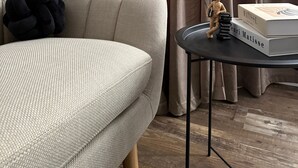 Ein Wohnzimmer mit einem beigefarbenen Sofa, einem schwarzen Kissen, einem kleinen Tisch mit einem Buch und einer Figur sowie einem Fenster mit Vorhängen.