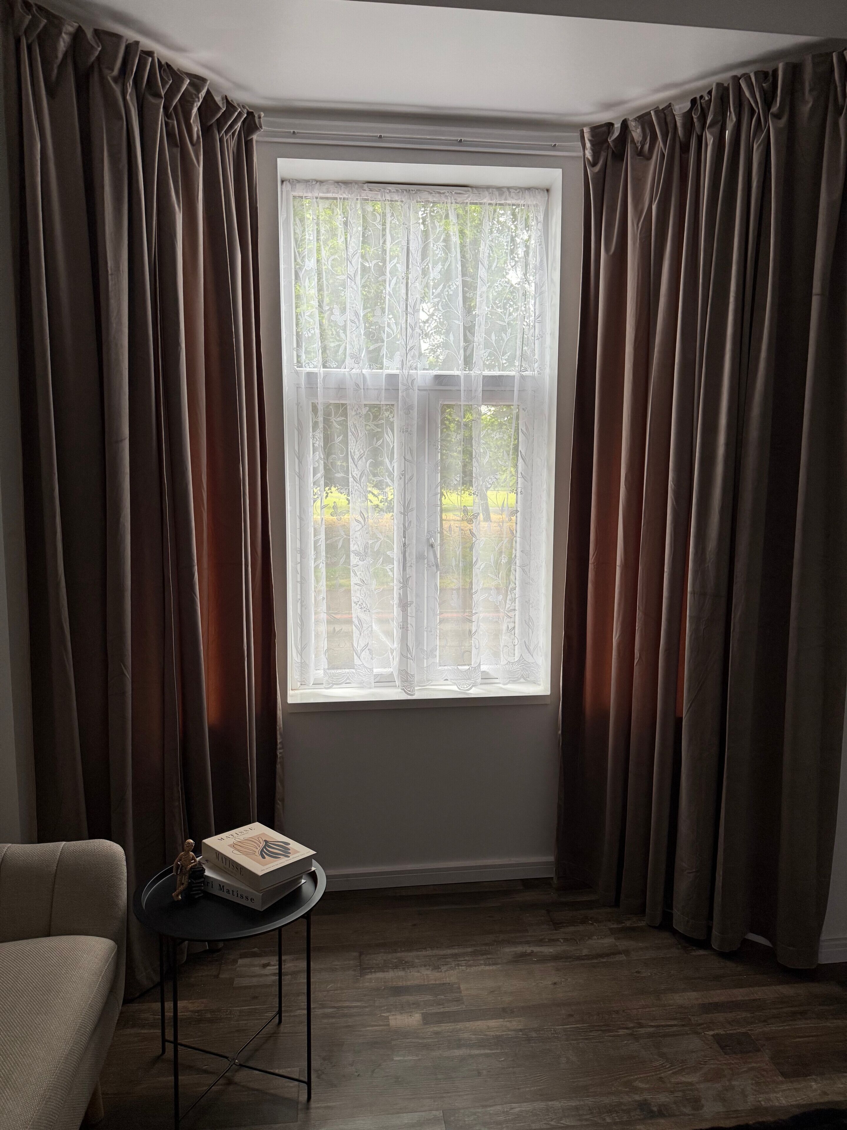 Una habitación con un ventanal grande, cortinas oscuras, una mesita con una caja y una silla.