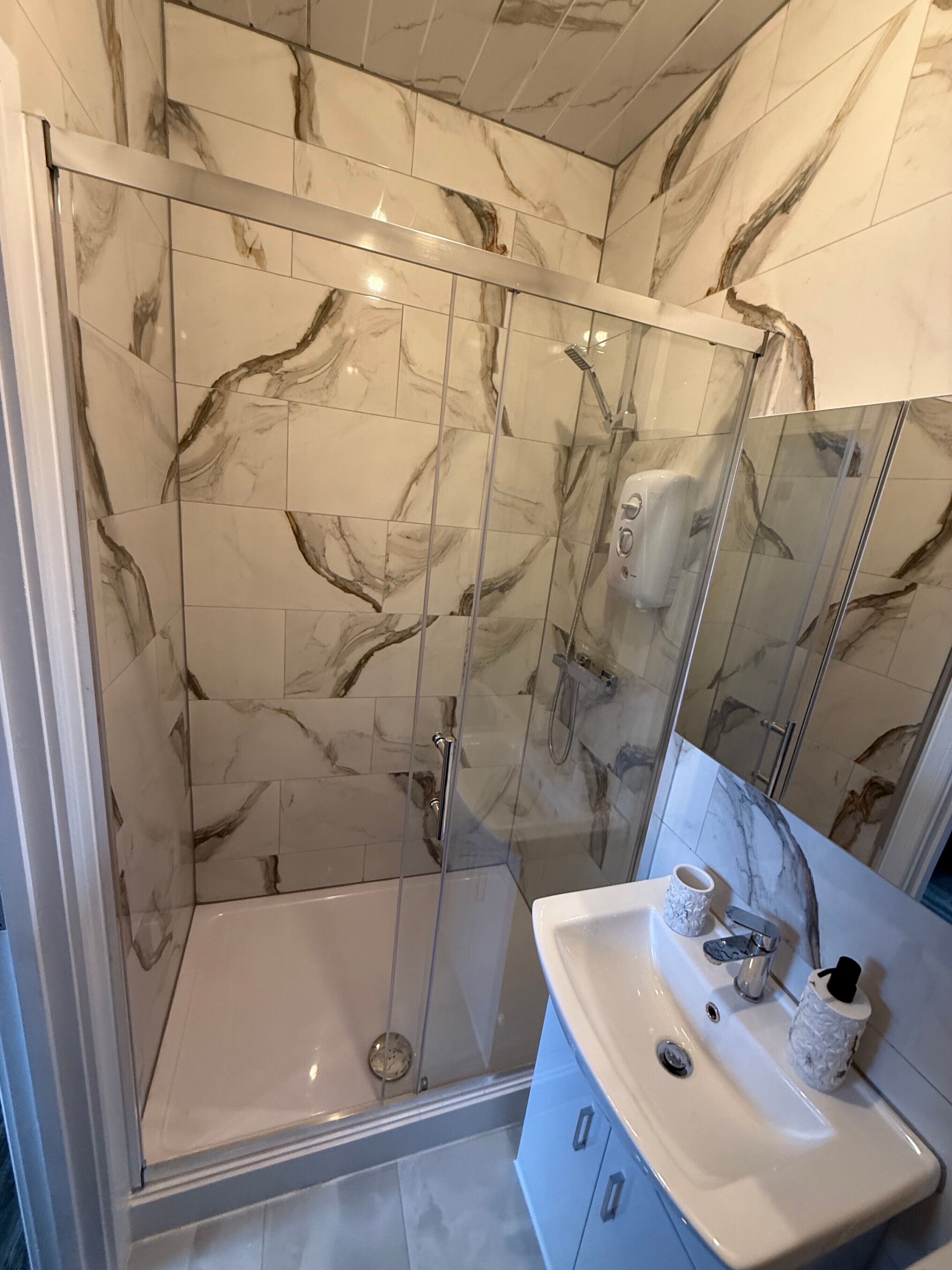 Un baño con ducha de vidrio, azulejos de mármol, un lavamanos con espejo y un portavasos.