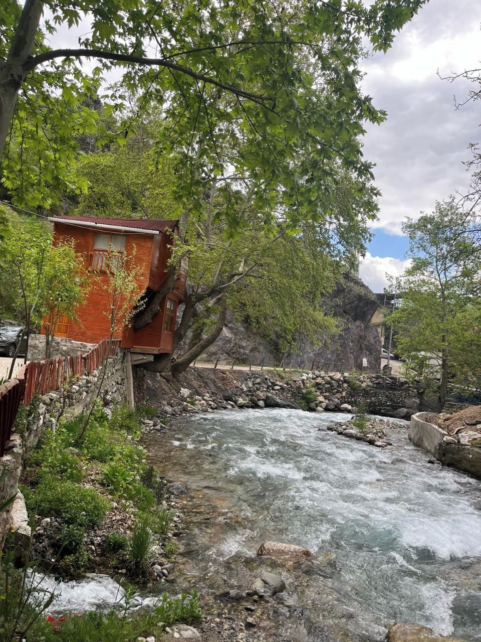 Une cabane en bois rustique, avec des fondations en pierre, située au bord d’une rivière à courant rapide, et entourée d’une végétation luxuriante et d’arbres.