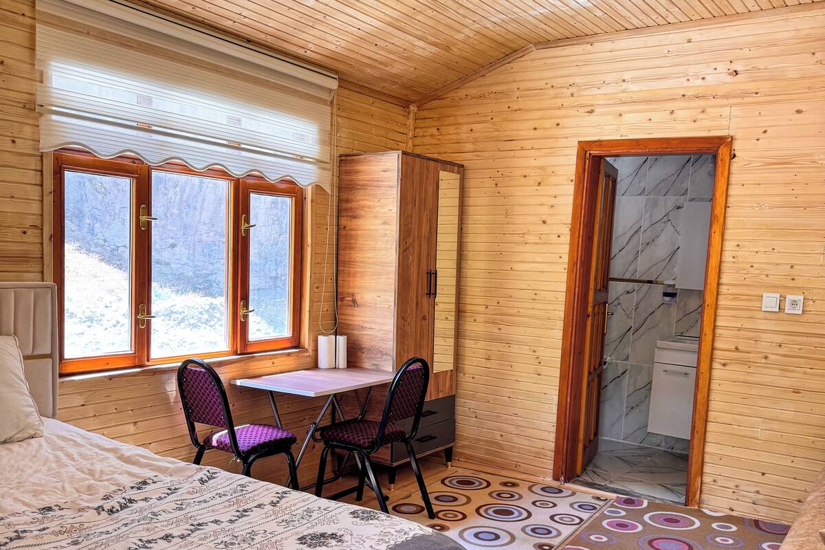 Une pièce en bois avec un lit, un bureau et une chaise. Il y a une fenêtre donnant sur des arbres et une porte menant à une salle de bain.