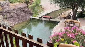 A rustic outdoor setting with a small pond, wooden deck, picnic table, and a brick building in the background. - Kapuzbaşı Baştürk Tesisi (Yahyali)