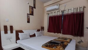 A bedroom with a bed, white pillows, a patterned blanket, a wall-mounted air conditioner, and a window with red curtains. - Hotel Natraj (Omkareshwar)