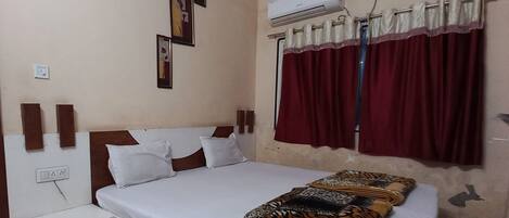 A bedroom with a bed, white pillows, a patterned blanket, a wall-mounted air conditioner, and a window with red curtains.