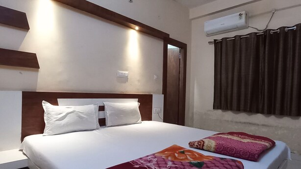 A hotel room with a large bed, a wooden headboard, a window with curtains, and an air conditioning unit on the wall. - Hotel Natraj (Omkareshwar)