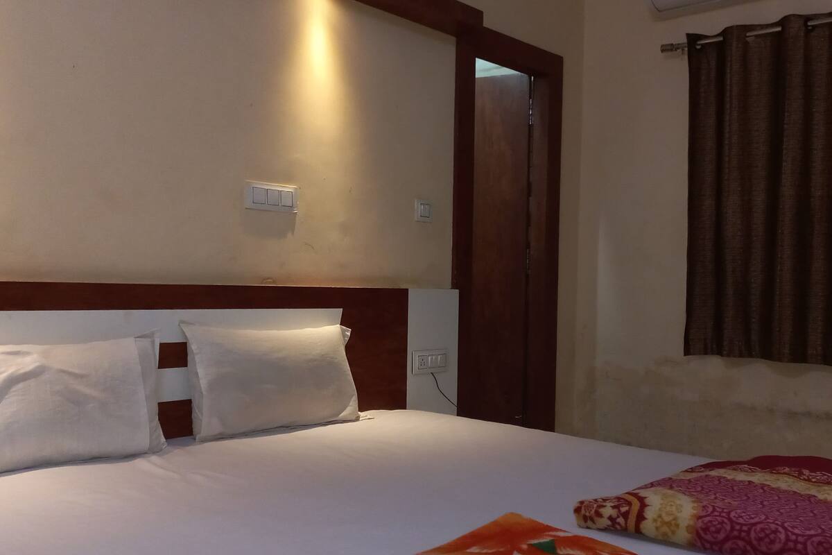 A hotel room with a bed, white pillows, a wooden headboard, a window with curtains, and a door.