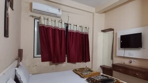 A hotel room with a bed, a flat-screen TV mounted on the wall, an air conditioning unit, and a window with red curtains. - Hotel Natraj (Omkareshwar)