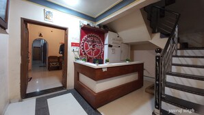 A reception desk with a wooden finish and a red decorative panel, a staircase with metal railings, and a hallway leading to another room. - Hotel Natraj (Omkareshwar)
