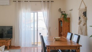 A dining area with a wooden table and chairs, a television on a stand, a cabinet with a plant, and a sliding glass door with curtains. - Sunny and Spacious Apartment with Balcony! Walk to Beach & Town (Lagos)