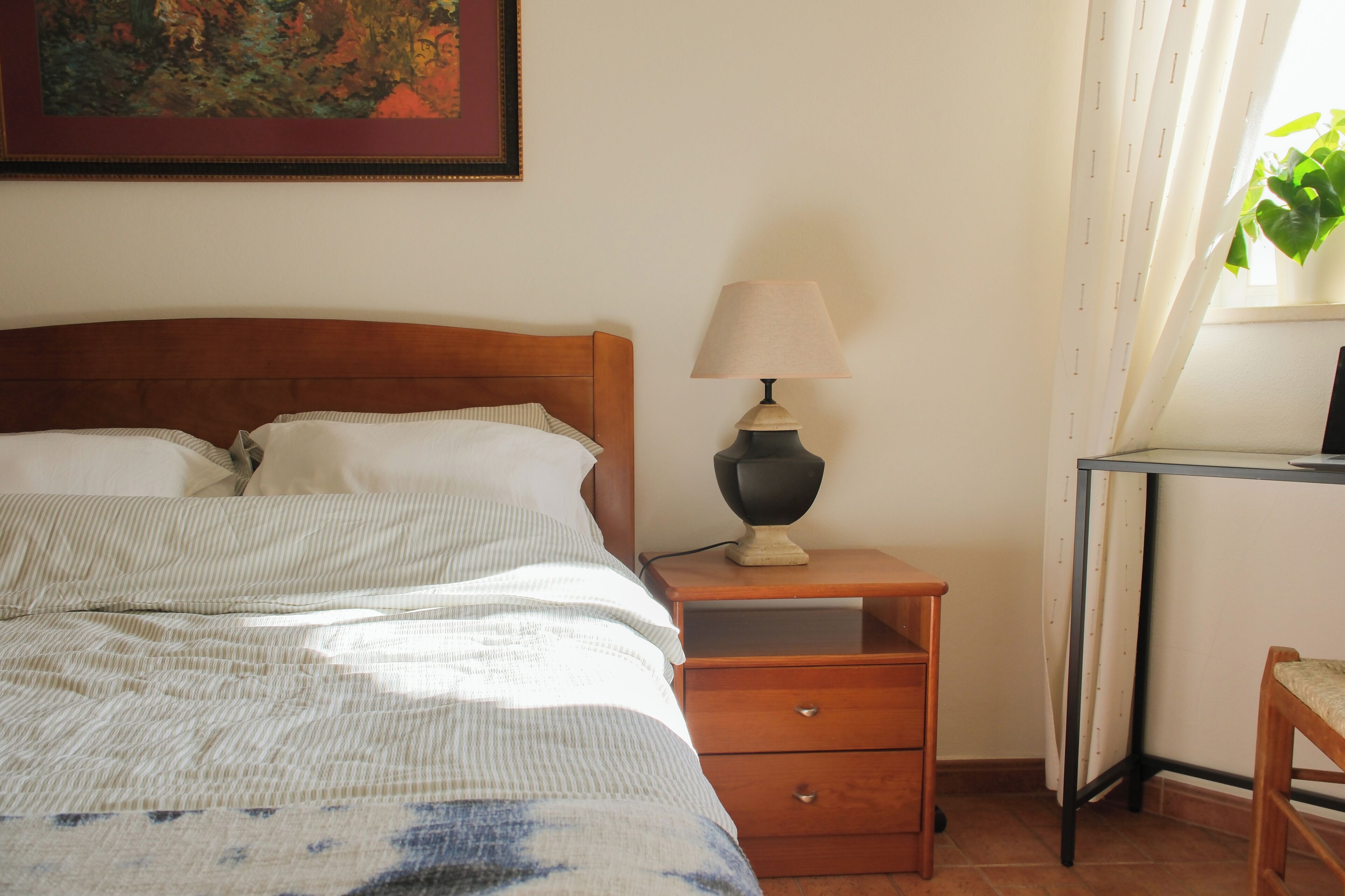 Una cama bien hecha con un edredón a rayas, una mesita de noche con lámpara, una pequeña mesita auxiliar de madera y una silla.