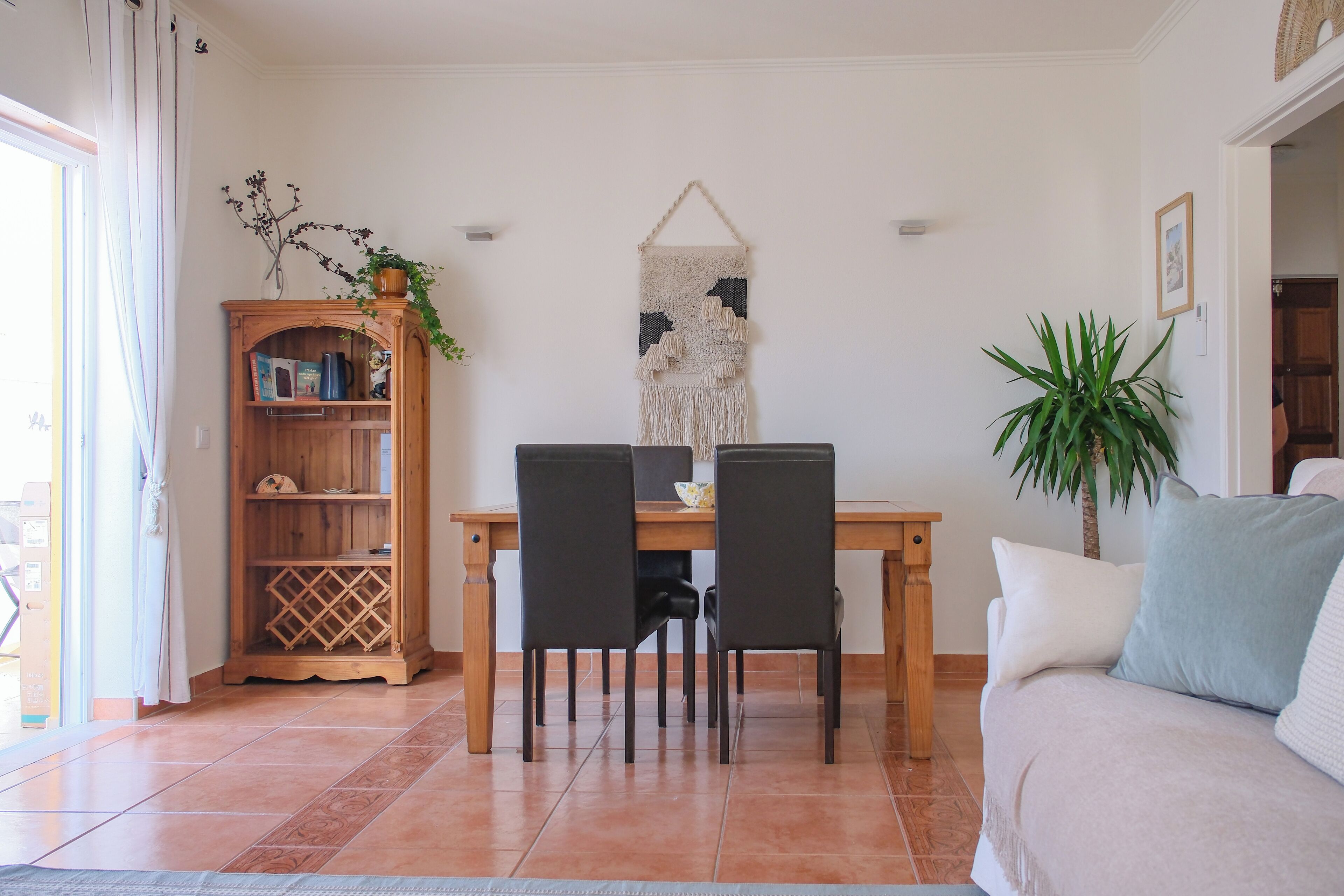 Un coin repas avec une table en bois, deux chaises, une bibliothèque, une décoration murale en macramé et une plante en pot.