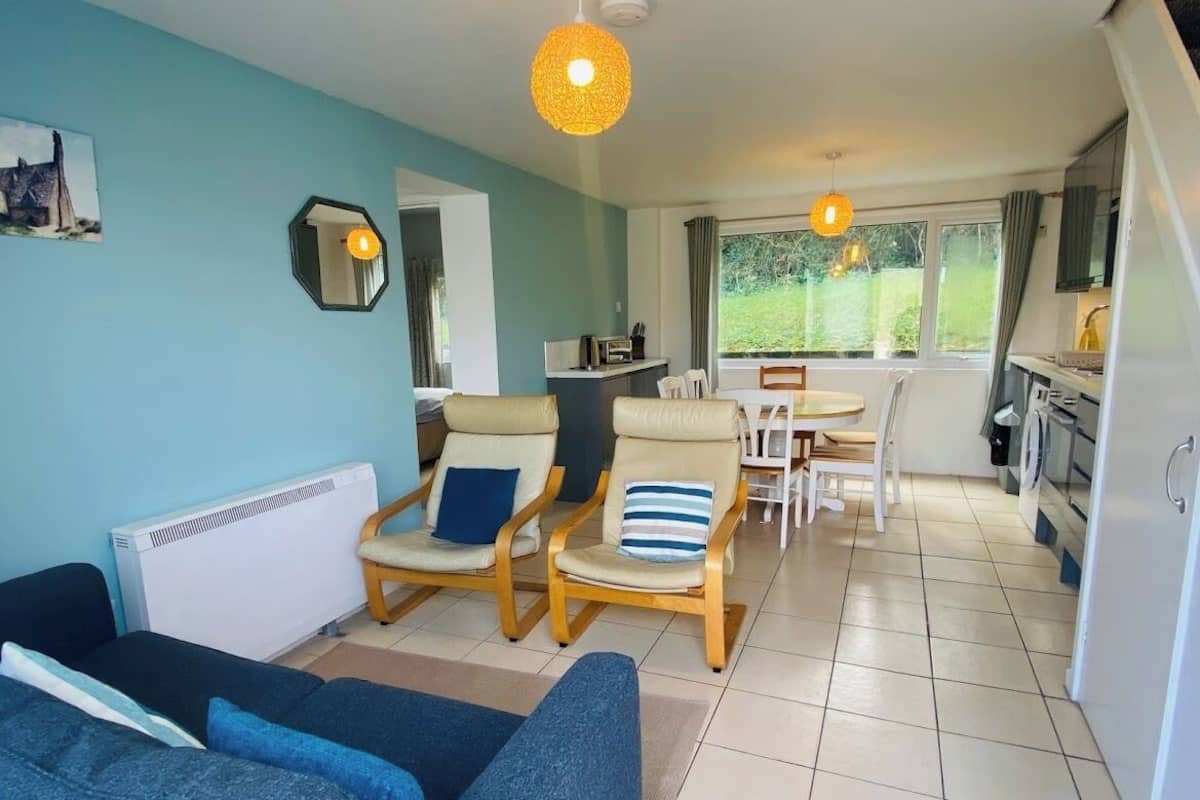 A living room with a sofa, two armchairs, a dining table with chairs, a kitchen area, and a large window with a view of greenery.
