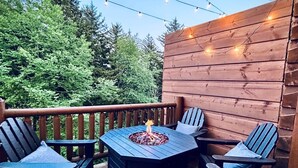 Eine Holzterrasse mit einem sechseckigen Tisch, umgeben von Stühlen, Lichterketten darüber und einer Feuerstelle in der Mitte.