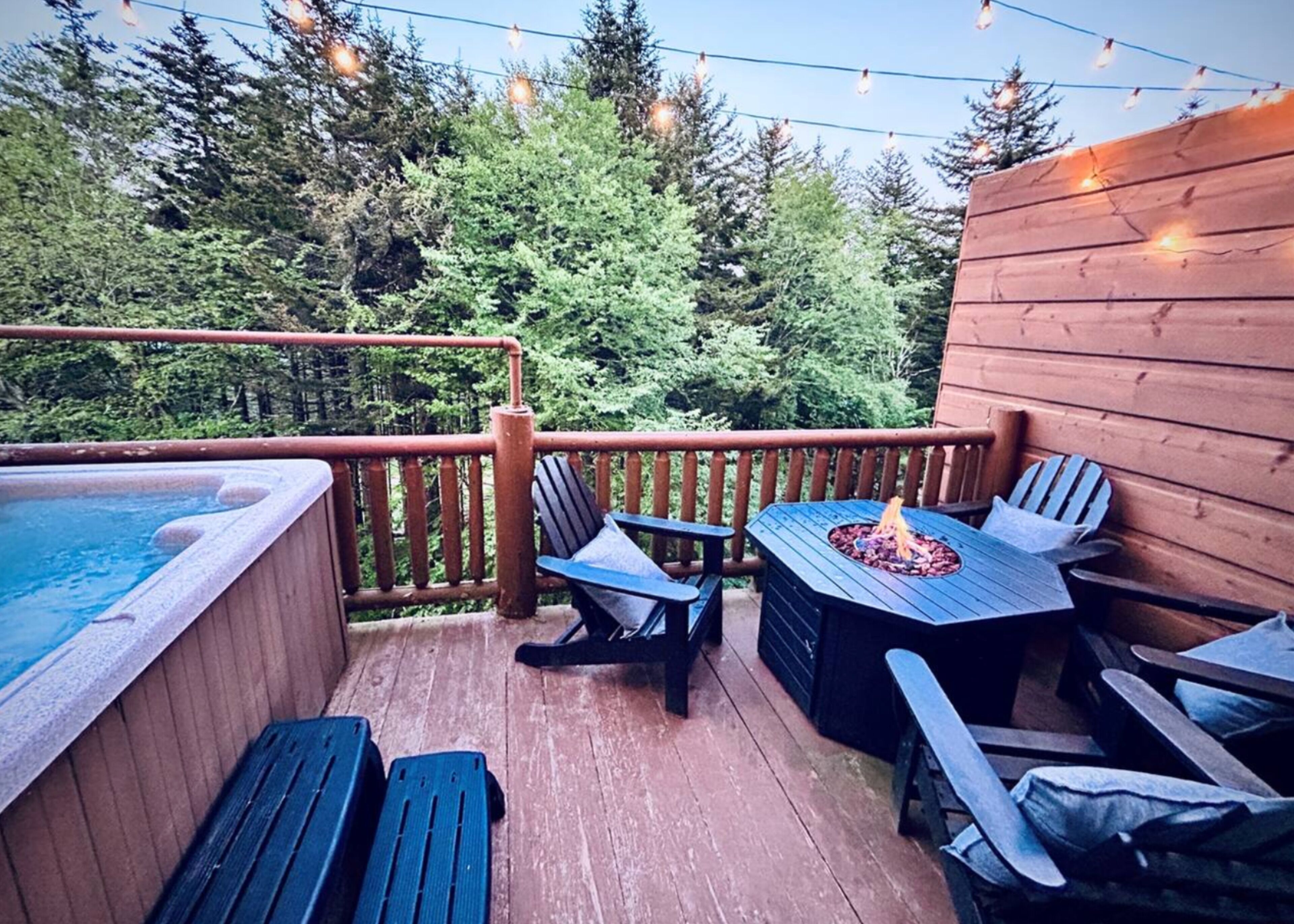 An outdoor deck with a hot tub, wooden deck chairs, a table with a fire pit, and string lights.