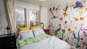Un dormitorio con cama, cortinas, cómoda y un cuadro floral en la pared.