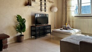 Un salon avec un téléviseur posé sur un meuble noir, une plante en pot, une table basse ronde avec une télécommande, et une fenêtre avec des rideaux.