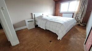 Une chambre avec un plancher en bois, un lit avec une literie blanche, une table de chevet et une fenêtre avec des rideaux.