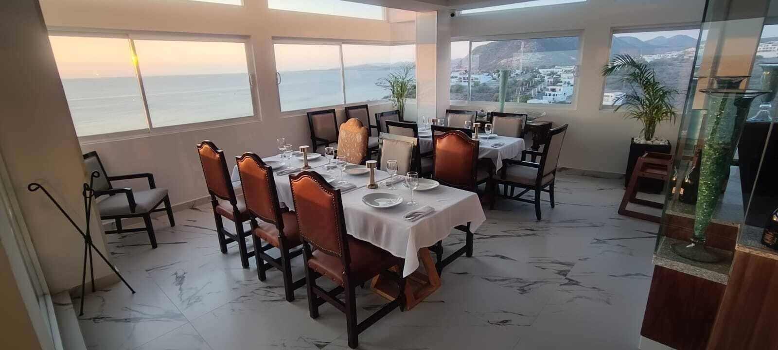 Un comedor con una mesa dispuesta para cuatro, ventanas amplias con vista al mar y una planta en un pedestal.