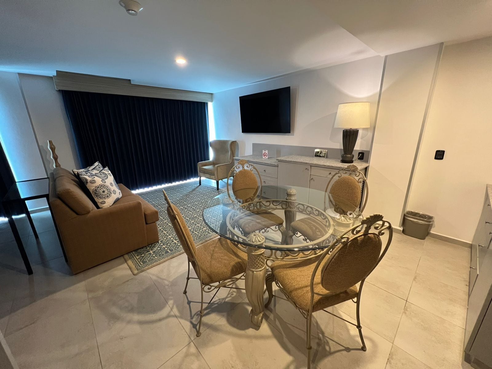 A modern living room with a glass-top dining table, cushioned chairs, a brown sofa, and a large window with dark curtains.